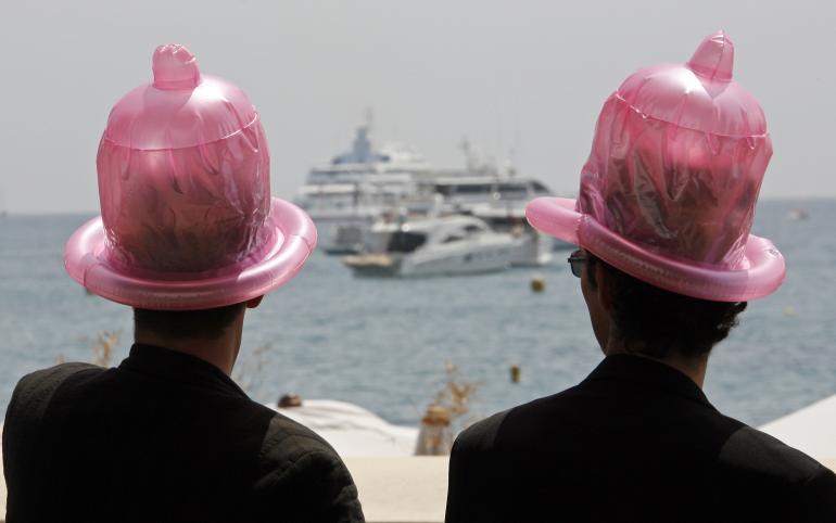 Photo de deux hommes avec un chapeau en forme de préservatif sur la tête et qui regardent la mer et un bateau