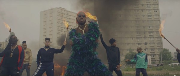 Kiddy Smile casquette rouge à l'envers et lunettes de soleil, croise les bras dans une posture lascive, vêtu d'un vêtement en froufrou vert. Arrière-plan son crew vêtu de noir porte des torches enflammées.